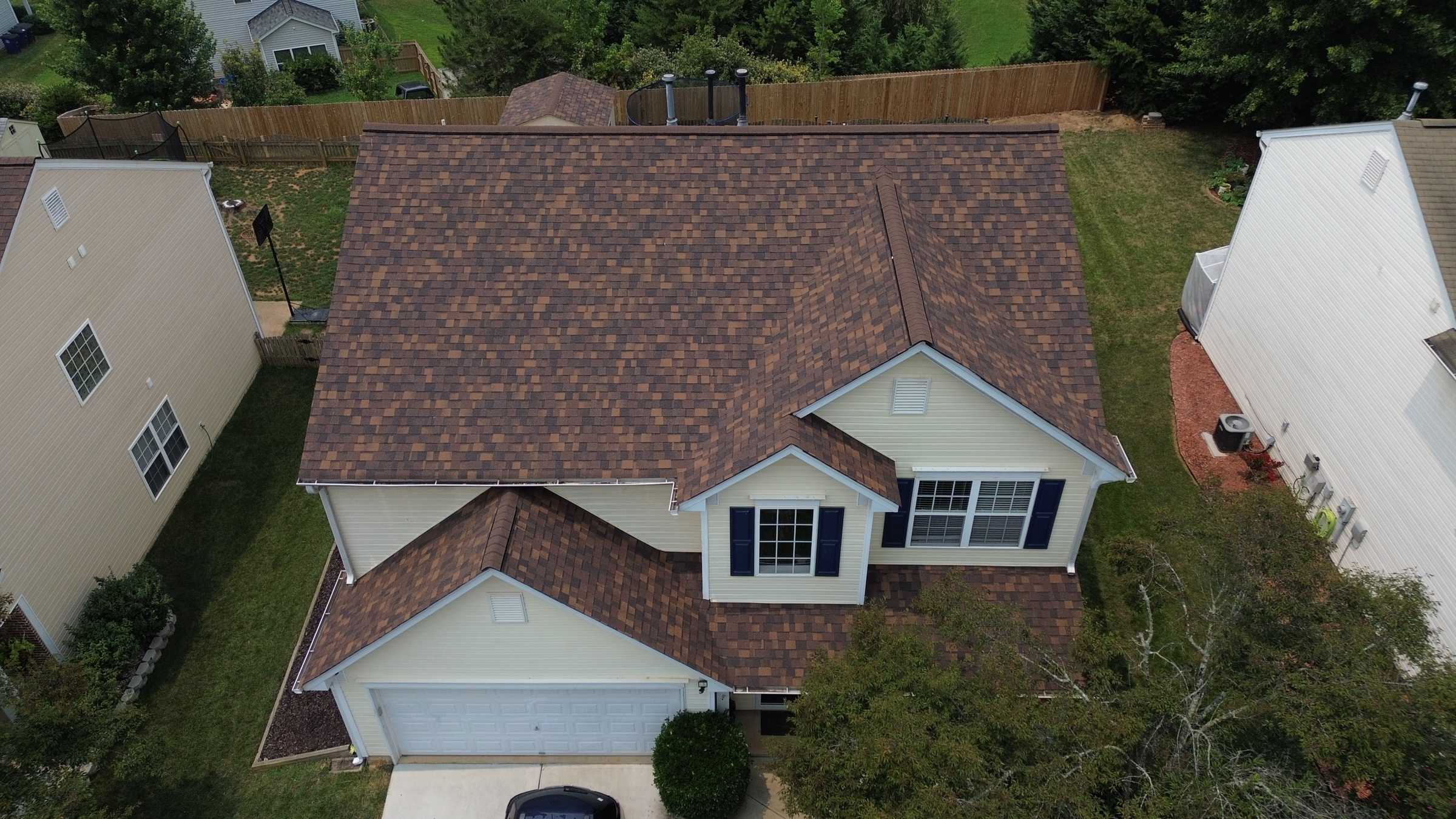 Drone view of a two-story house with a new brown shingle roof between neighboring houses.