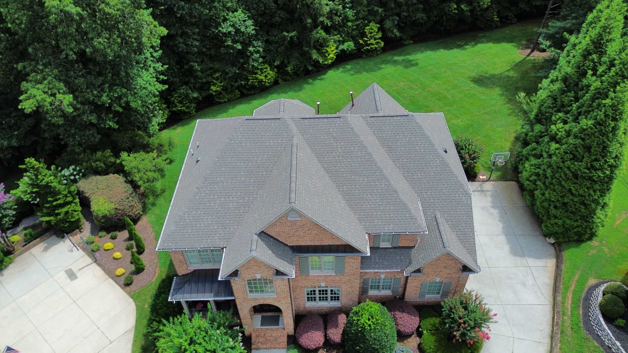 Drone view of a large brick house with a new gray shingle roof and a wide lawn behind it.